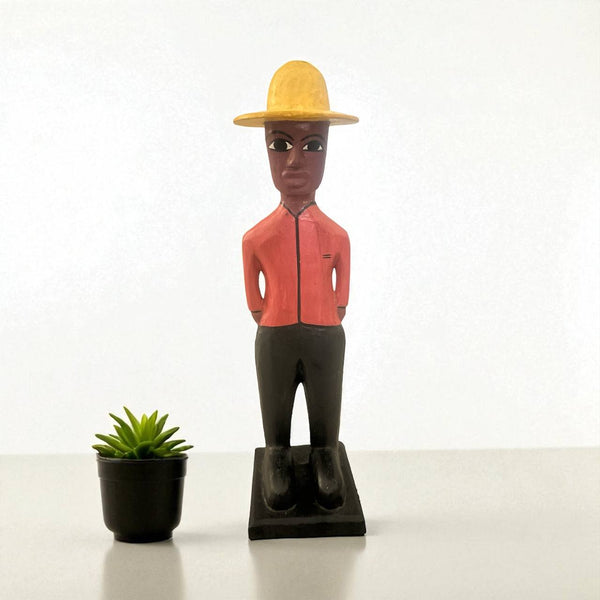 Wooden figurine of a person wearing a yellow hat and red shirt next to a small potted plant on a white background