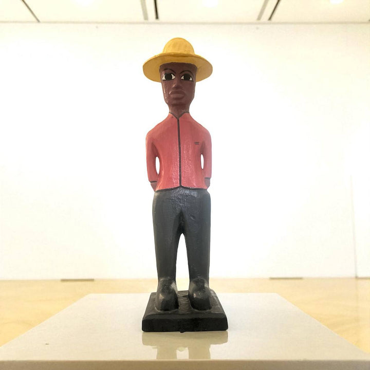 Wooden figurine of a person wearing a yellow hat and red jacket on a white surface.