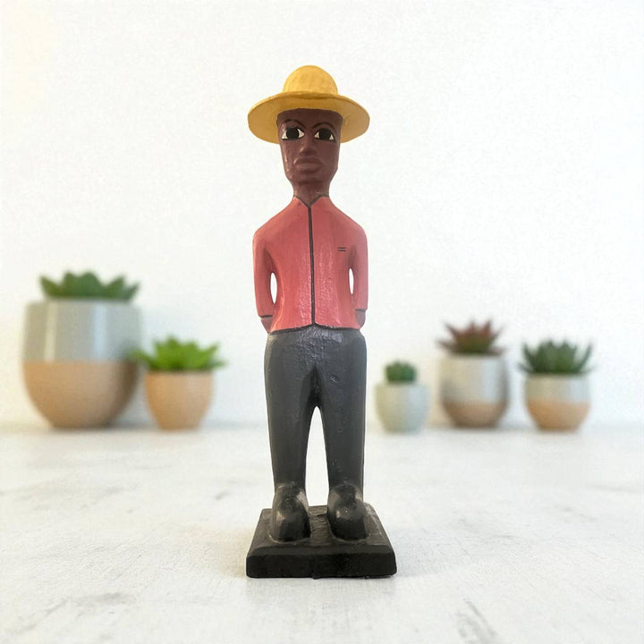 Figurine of a person wearing a yellow hat and pink shirt with potted plants in the background.