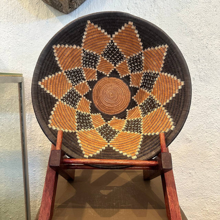 Decorative woven basket with geometric pattern on a wooden stand against a textured wall.