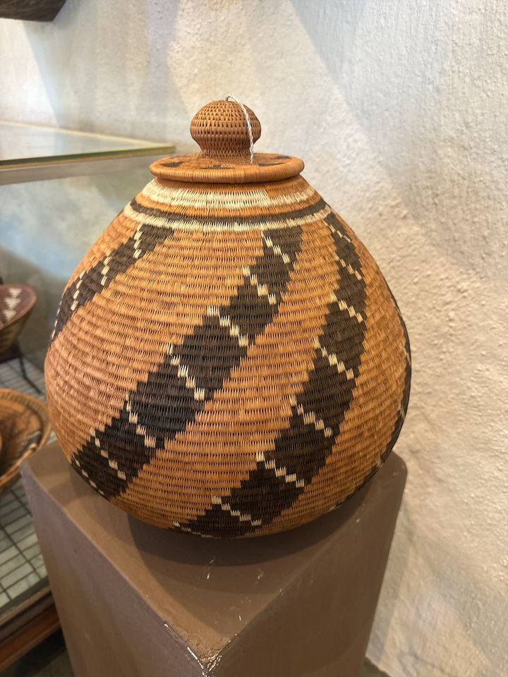 Woven basket with a lid on a pedestal against a textured wall.